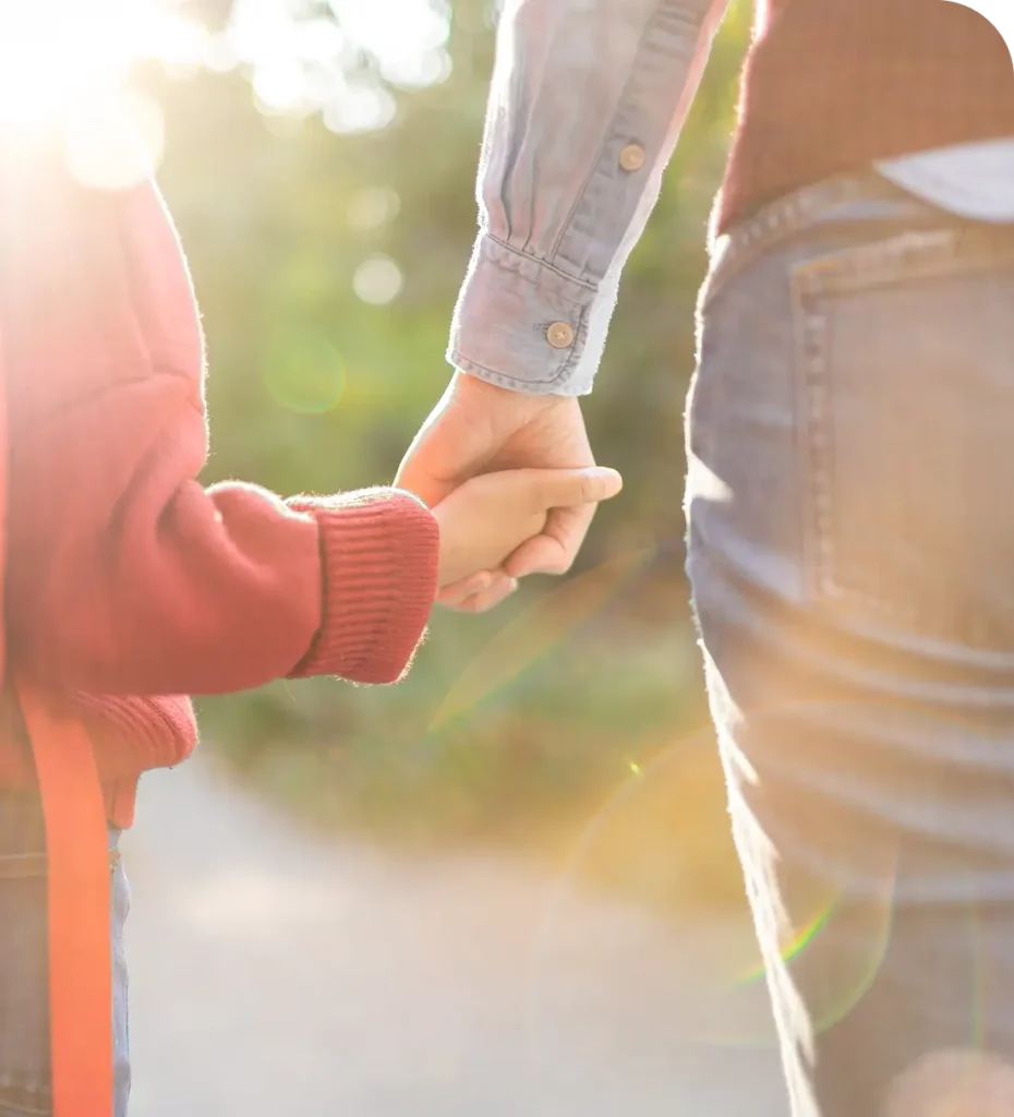 Nahaufnahme von zwei Personen, die sich an den Händen halten, links ein Kind mit rotem Pullover, rechts ein Erwachsener mit Jeans und hellblauem Hemd.