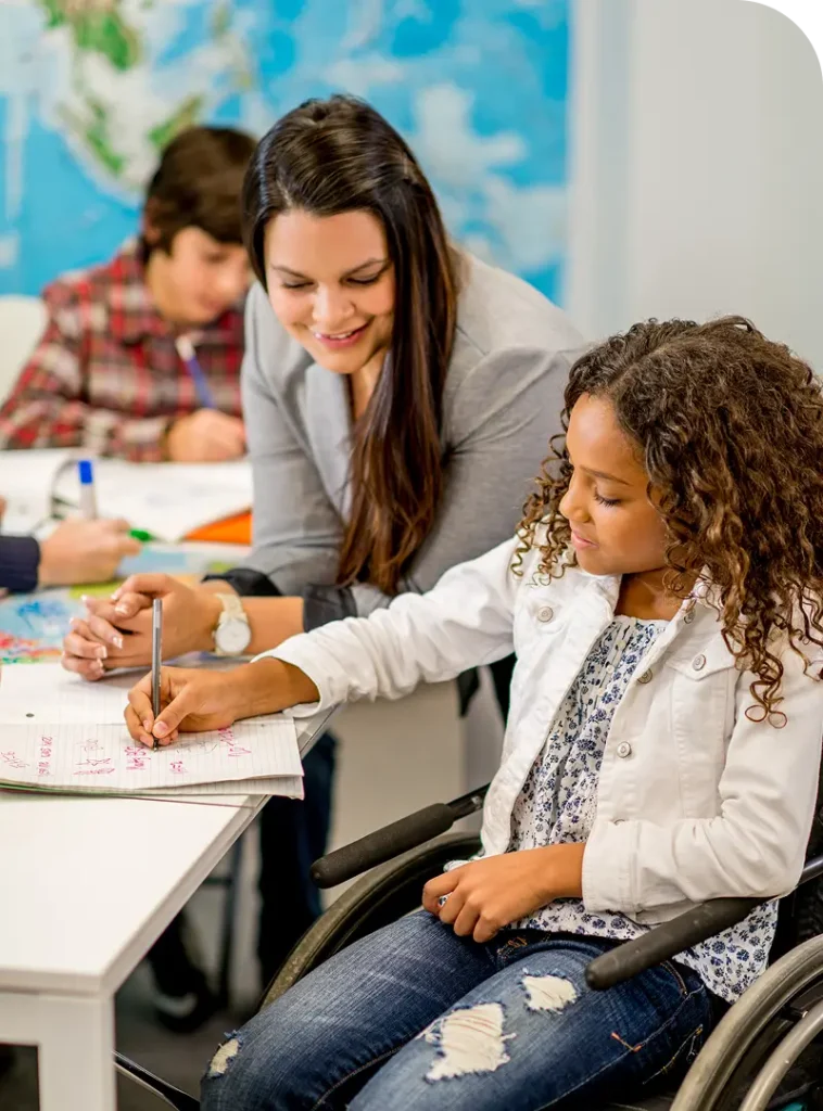 Kind im Rollstuhl schreibt an einem Tisch mit Unterstützung einer erwachsenen Person im Klassenzimmer.