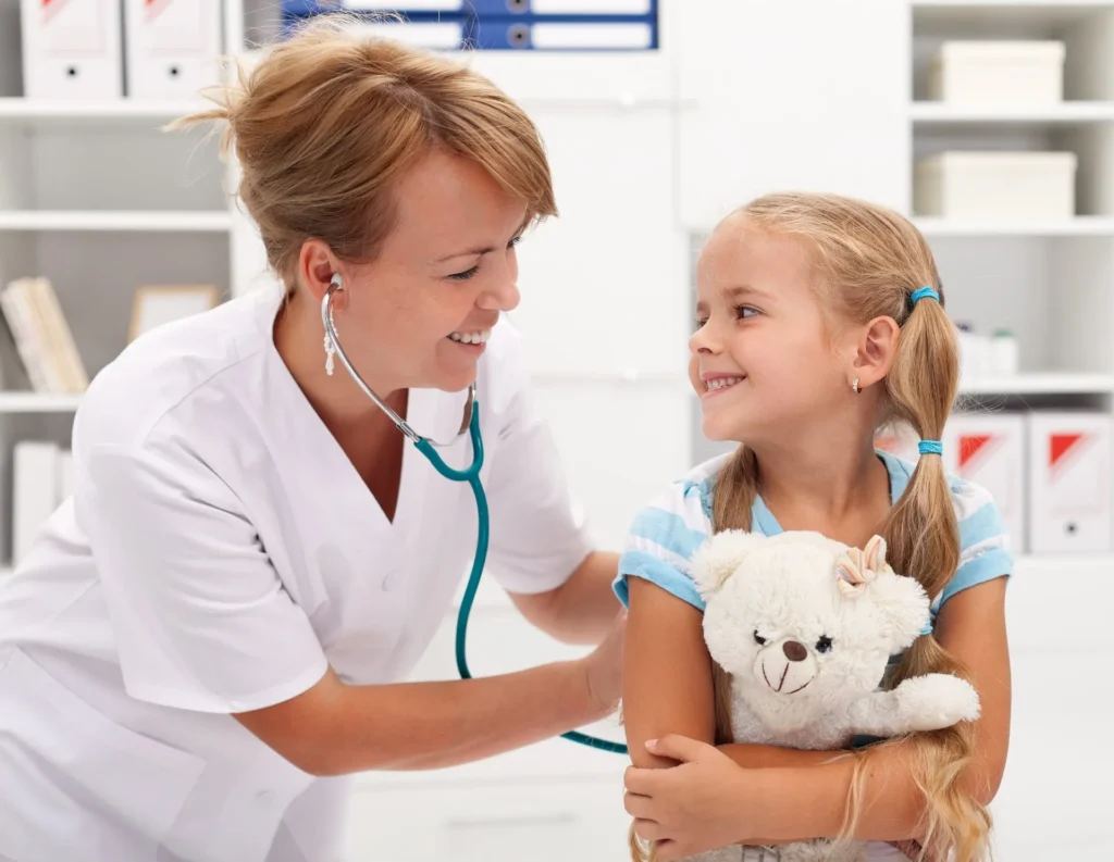 Ärztin mit Stethoskop hört Mädchen mit Zöpfen ab, das einen Teddybär hält